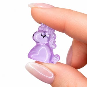 May include: A small, translucent purple unicorn figurine crafted from a smooth, glossy material. The unicorn features a stylized mane, a curled tail, and simple black painted lines to represent closed eyes and eyelashes on its side profile.