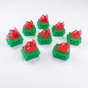 May include: A collection of nine green and red decorative keycaps. Each keycap features a green square base with a smiling face and a translucent red flame design on top. The keycaps are arranged on a white surface.