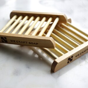 Op de afbeelding: Twee houten zeepbakjes met een lamellenontwerp. De bakjes zijn op elkaar gestapeld. Het onderste bakje heeft de tekst "WE CRAFT SOAP" gegraveerd op de voorkant.