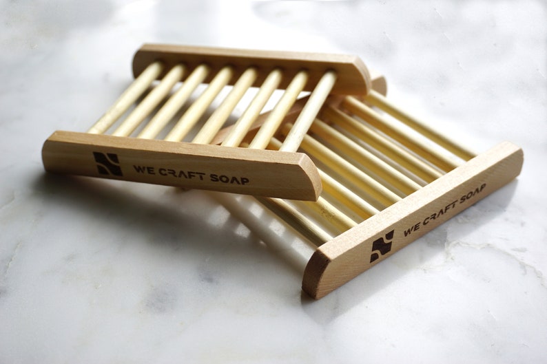Op de afbeelding: Twee houten zeepbakjes met een lamellenontwerp. De bakjes zijn op elkaar gestapeld. Het onderste bakje heeft de tekst "WE CRAFT SOAP" gegraveerd op de voorkant.