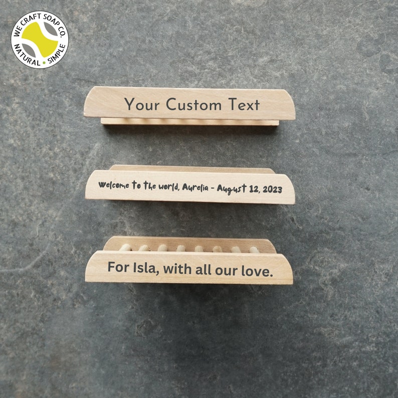 Op de afbeelding: Een houten zeepbakje met een natuurlijke afwerking. Het zeepbakje heeft een verhoogde rand en een decoratief ontwerp. Het zeepbakje is gegraveerd met de tekst "Your Custom Text", "Welcome to the world, Aurelia - August 12, 2023" en "For Isla, with all our love."