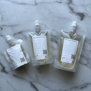 May include: Three clear plastic pouches filled with a light yellow liquid. Each pouch has a white cap and a label that reads "100% Organic Coconut Oil".
