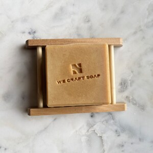 Op de afbeelding: Een reep natuurlijke zeep in een houten zeephouder. De zeep is lichtbruin van kleur en heeft de woorden "WE CRAFT SOAP" erop gegraveerd.