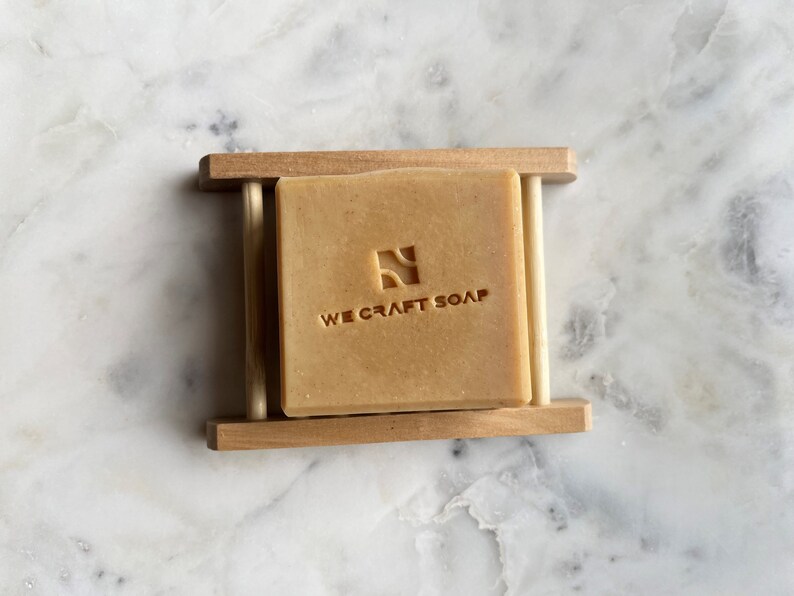 Op de afbeelding: Een reep natuurlijke zeep in een houten zeephouder. De zeep is lichtbruin van kleur en heeft de woorden "WE CRAFT SOAP" erop gegraveerd.