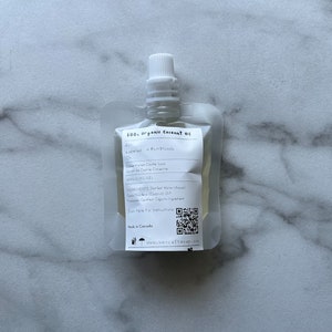 May include: A white pouch with a clear window, containing a 200 ml bottle of organic coconut oil. The pouch has a label with the text "200 ml organic coconut oil" and a QR code. The label also lists the ingredients: Distilled Water (Aqua), Cocos Nucifera (Coconut) Oil, Sodium Citrate, Citric Acid.