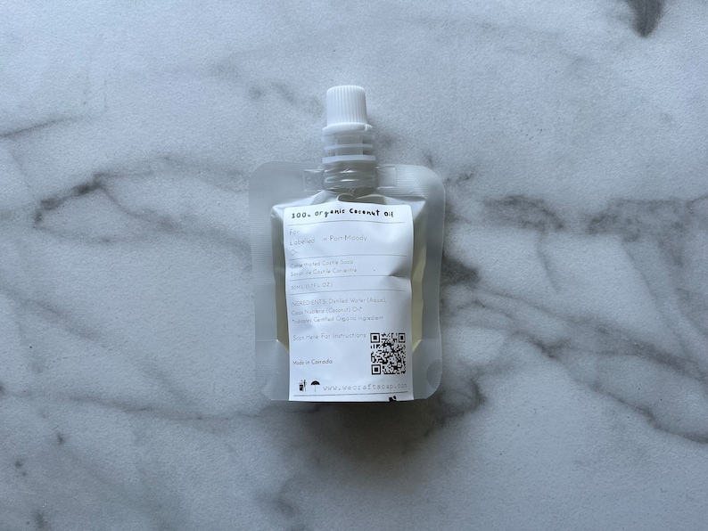 May include: A white pouch with a clear window, containing a 200 ml bottle of organic coconut oil. The pouch has a label with the text "200 ml organic coconut oil" and a QR code. The label also lists the ingredients: Distilled Water (Aqua), Cocos Nucifera (Coconut) Oil, Sodium Citrate, Citric Acid.