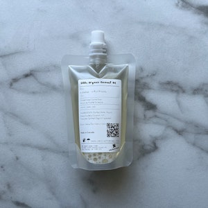 May include: A clear plastic pouch filled with a light yellow liquid. The pouch has a white label with the text "100% Organic Coconut Oil" and other information about the product. The pouch is on a white marble surface.
