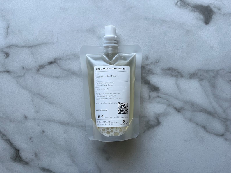 May include: A clear plastic pouch filled with a light yellow liquid. The pouch has a white label with the text "100% Organic Coconut Oil" and other information about the product. The pouch is on a white marble surface.