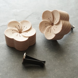Puede incluir: Dos ambientadores de coche de madera con forma de flor de cerezo. Los ambientadores son de color marrón claro y tienen un diseño detallado de una flor con un agujero central. También se incluye una pinza para ambientadores de coche negra.