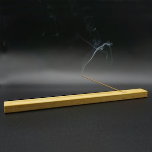Könnte beinhalten: Ein hölzerner Räucherstäbchenhalter mit einem brennenden Räucherstäbchen, das Rauch abgibt. Der Halter ist aus hellbraunem Holz und hat eine rechteckige Form.