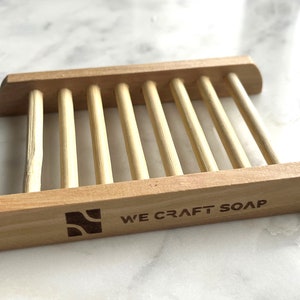 Op de afbeelding: Houten zeepbakje met een lamellenontwerp en de woorden "WE CRAFT SOAP" gegraveerd op de voorkant.