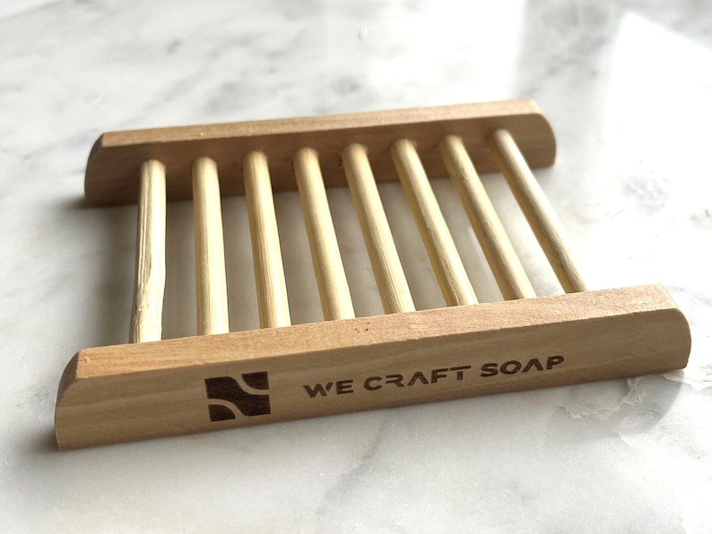 Op de afbeelding: Houten zeepbakje met een lamellenontwerp en de woorden "WE CRAFT SOAP" gegraveerd op de voorkant.
