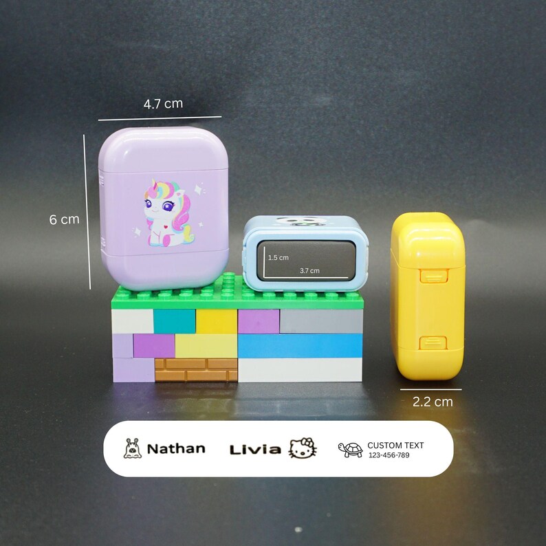 May include: A collection of three small, colorful items. A lavender case (6 cm tall, 4.7 cm wide) with a unicorn graphic, a blue rectangular item (3.7 cm wide, 1.5 cm tall), and a yellow item (2.2 cm wide). A white label with text and icons is at the bottom.