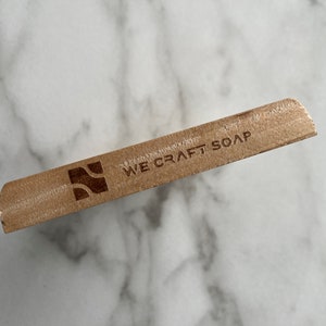 Op de afbeelding: Een houten zeepstaaf met de tekst "WE CRAFT SOAP" en een gestileerd golflogo.