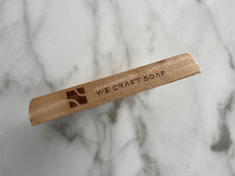 Op de afbeelding: Een houten zeepstaaf met de tekst "WE CRAFT SOAP" en een gestileerd golflogo.