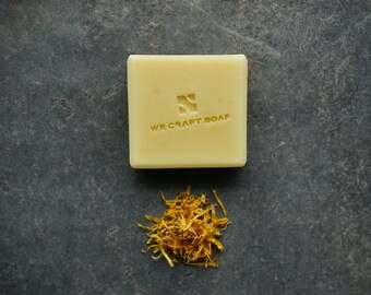 Könnte beinhalten: Eine Stange hellgelber Seife mit den Worten "We Craft Soap" und einem geprägten Logo auf der Vorderseite. Unter der Seife befindet sich ein kleiner Haufen getrockneter gelber Blütenblätter.