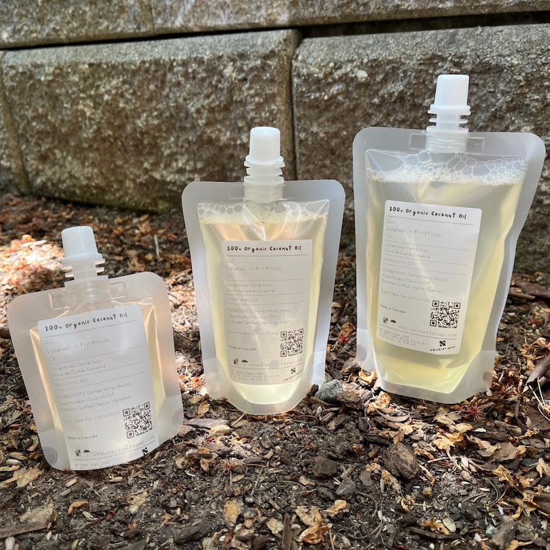 May include: Three clear plastic pouches filled with a light yellow liquid. The pouches are labeled "100% Organic Coconut Oil" and have a QR code. The pouches are sitting on a bed of brown mulch.