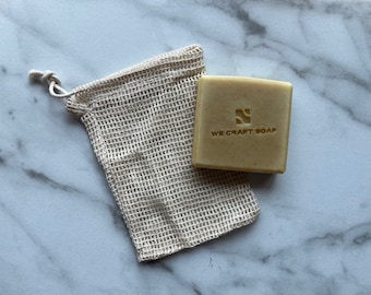 May include: A bar of soap in a beige mesh bag. The soap is a light yellow color and has the text "WE CRAFT SOAP" printed on it.