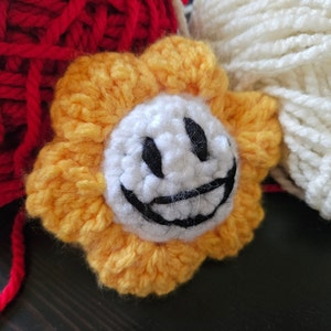 May include: A crocheted yellow flower with a white face and a black smiling mouth. The flower is surrounded by red and white yarn.