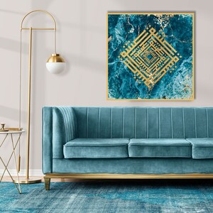 Op de afbeelding: Een tealblauwe fluwelen bank met gouden accenten staat voor een muur met een vierkant canvasprint. De canvasprint heeft een gouden Arabisch kalligrafieontwerp op een blauwe en gouden marmerachtergrond.