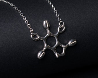 May include: A silver necklace with a molecular structure pendant. The pendant is a stylized representation of a molecule, with a central ring and six arms extending outward. The necklace is on a black background.