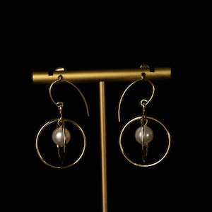 May include: Gold-coloured hoop earrings, each with a white pearl at the centre. The earrings are displayed on a gold stand against a black backdrop. The design is simple and elegant.