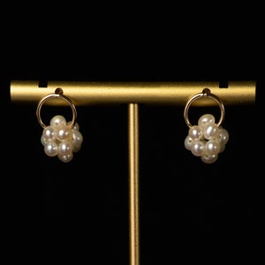 Peut inclure: Une paire de boucles d'oreilles cr&eacute;oles en or avec un groupe de perles blanches suspendues &agrave; chaque cr&eacute;ole. Les boucles d'oreilles sont pr&eacute;sent&eacute;es sur un support en or sur un fond noir.