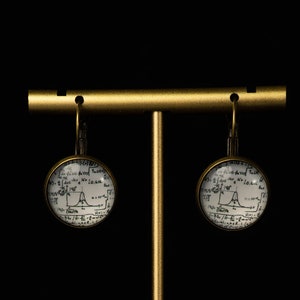 Puede incluir: Un par de pendientes de tono dorado con un colgante circular de vidrio blanco que presenta un diagrama científico en blanco y negro. Los pendientes se muestran en un soporte dorado sobre un fondo negro.