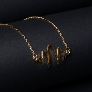 Puede incluir: Un collar de oro con un colgante ondulado de diseño abstracto. El collar está sobre un fondo negro.