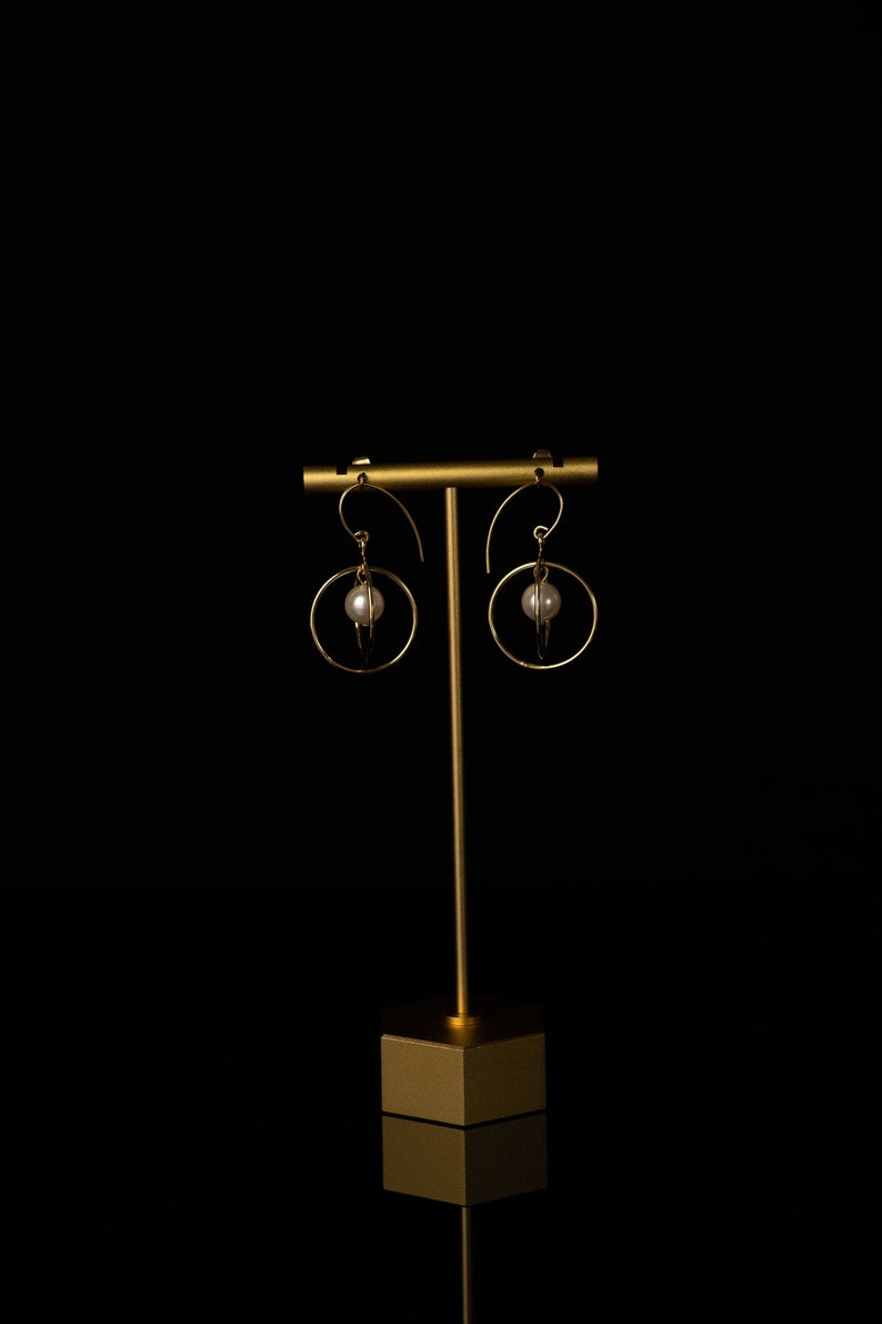 May include: Gold-coloured hoop earrings, each with a white pearl at the centre. The earrings are displayed on a gold stand against a black backdrop. The design is simple and elegant.