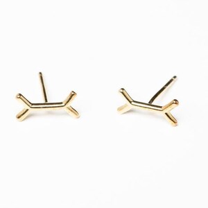 May include: A pair of gold-toned earrings with a simple, geometric design. Each earring features a small, abstract shape that resembles a branch or a stylized letter Y.