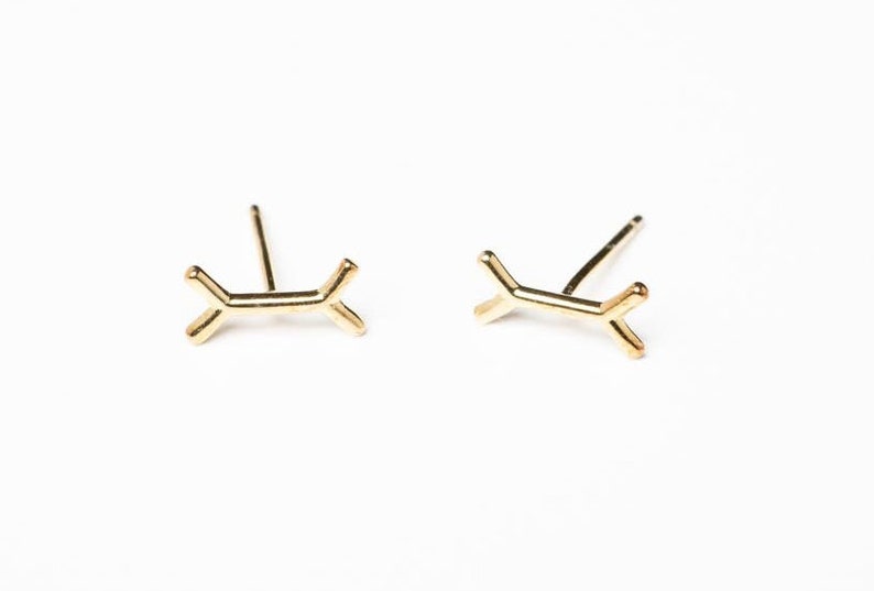 May include: A pair of gold-toned earrings with a simple, geometric design. Each earring features a small, abstract shape that resembles a branch or a stylized letter Y.