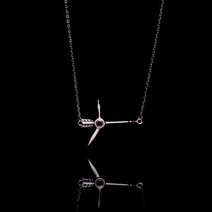 Puede incluir: Un collar de plata con una cadena delicada y un colgante con forma de flecha con un centro circular. El colgante está suspendido de la cadena y cuelga en el centro de la imagen.