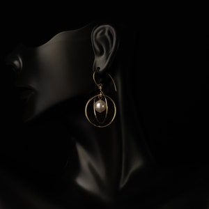 May include: Gold-coloured earrings with a pearl accent. The earrings feature a circular design with a dangling pearl in the centre. The jewellery is displayed on a black mannequin head.