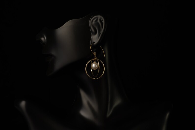 May include: Gold-coloured earrings with a pearl accent. The earrings feature a circular design with a dangling pearl in the centre. The jewellery is displayed on a black mannequin head.