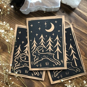 Peut inclure: Trois carnets marrons avec des illustrations à l'encre noire de lapins courant dans une forêt sous une lune croissante et des étoiles.