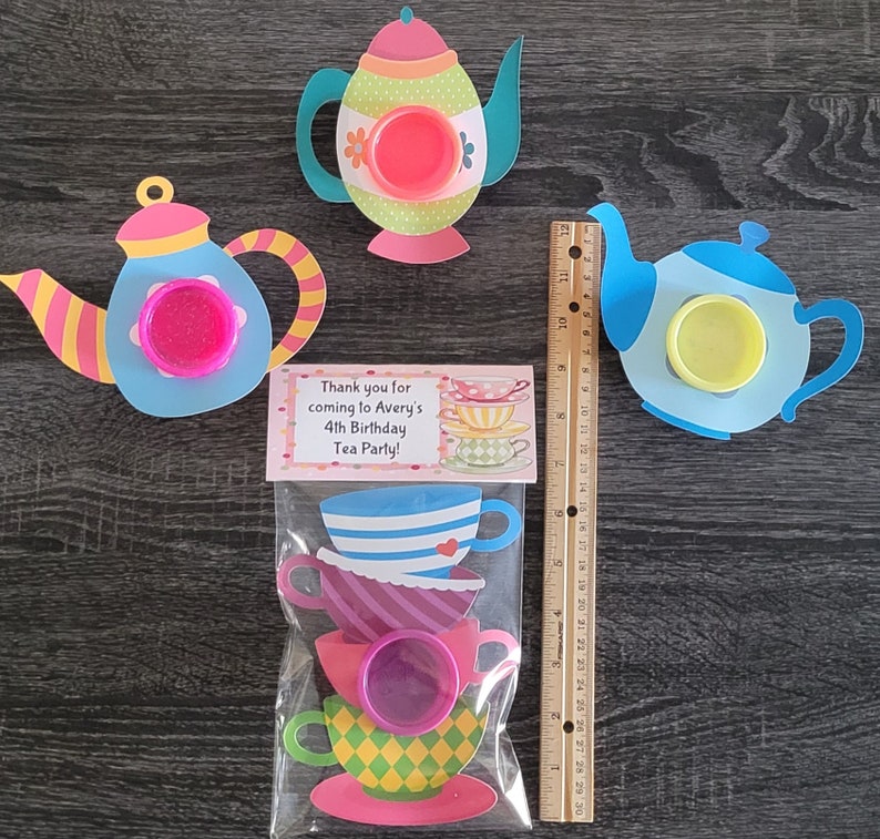 May include: A set of tea party favor bags with a thank you tag that reads "Thank you for coming to Avery's 4th Birthday Tea Party!" The bags contain a stack of teacups with a pink lid and a blue teapot with a pink lid.