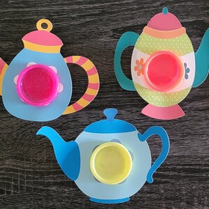 May include: Three colorful paper teapots with lids, each with a different colored lid and a different colored circle in the center. The teapots are on a dark wood surface.