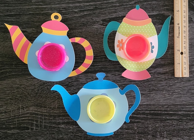 May include: Three colorful paper teapots with lids, each with a different colored lid and a different colored circle in the center. The teapots are on a dark wood surface.