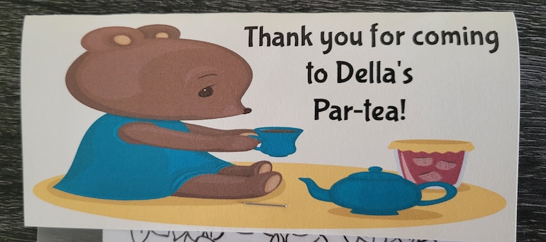 May include: Thank you for coming to Della's Par-tea! A brown bear wearing a blue dress sits at a table with a teapot and a cup of tea.