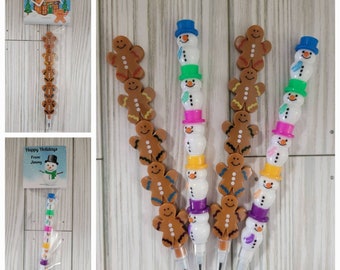 May include: Three sets of gingerbread man and snowman stackable pencil toppers. Each set has a gingerbread man with a smiling face and a snowman with a blue, green, pink, or yellow hat. The pencils are packaged in clear plastic bags with a "Happy Holidays" tag.