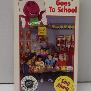 May include: A VHS video cassette cover featuring Barney the Dinosaur, a purple dinosaur with a green shirt, in a classroom setting. The cover reads "Barney Goes To School" and "Sing Along". The cover is for ages 2-8.