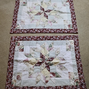May include: Two handmade quilted placemats. Each features a starburst design in floral and solid fabrics, with a burgundy floral border. The center is a mix of white, yellow, and pink floral patterns, set against a light green checkered background.