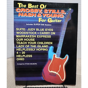 Könnte beinhalten: Ein Vintage-Gitarren-Notenbuch mit dem Titel "The Best Of Crosby, Stills, Nash & Young For Guitar". Das Cover zeigt eine stilisierte Gitarrengrafik in Rot, Blau und Weiß. Zu den Titeln gehören "Suite: Judy Blue Eyes" und "Woodstock".
