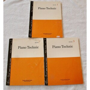 May include: Three vintage piano technique books. Each book has a cream-colored top half with the title "Piano Technic" and an orange bottom half. The spines are black with white text. The books are labeled "Book 1", "Book 2", and "Book 3".