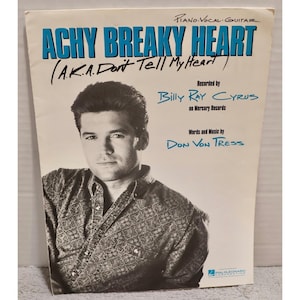 Puede incluir: Portada de partituras de "Achy Breaky Heart" de Billy Ray Cyrus. El título está en letras grandes y azules. La portada presenta un retrato de un hombre con una camisa estampada. El texto incluye el título alternativo de la canción, el artista y el compositor.