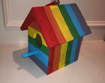 May include: A colorful, handmade birdhouse painted in a rainbow of colors. The birdhouse features a round entry hole, a blue perch, and a striped ribbon for hanging. The roof and sides are painted in vertical stripes of red, orange, yellow, green, blue, and purple.