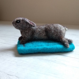 Op de afbeelding: Een bruin en grijs naaldgevoeld konijn dat op een turquoise blauw viltbed ligt.