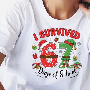 May include: White t-shirt with the text "I Survived 67 Days of School" in red, green, and black. The design includes a Santa hat, elf hat, presents, and books. The shirt is worn by a person.