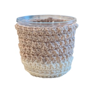 May include: A clear glass jar with a crocheted cozy. The cozy is two-toned, with a light tan color at the top that fades into a creamy white at the bottom. The cozy is made of thick, textured yarn.